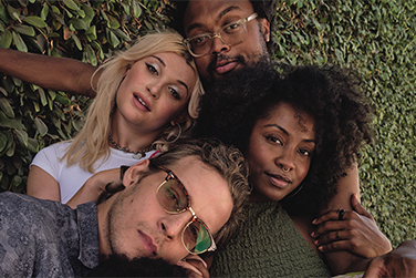A diverse group of four young adults—two men and two women—relax closely together in front of a leafy green wall, smiling and enjoying each other’s company.